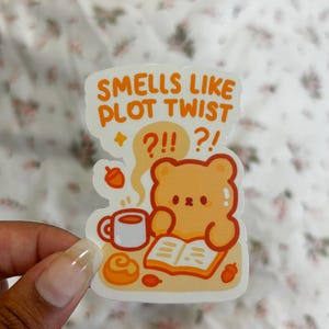 May include: A sticker featuring a cute bear reading a book with a cup of coffee and a pastry. The sticker has the text "Smells Like Plot Twist" with question marks. The bear is light brown with orange accents.