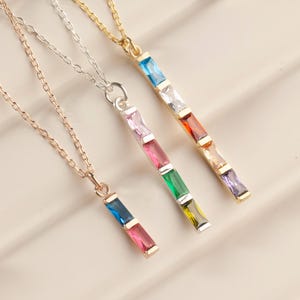 May include: Three delicate necklaces featuring vertical bar pendants set with rectangular multi-colored gemstones. One necklace is rose gold, one is silver, and one is gold, each with a fine chain and a series of vibrant, faceted stones.
