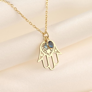 May include: Gold-toned necklace featuring a Hamsa hand pendant with intricate cut-out details. The pendant is adorned with blue gemstones and hangs from a delicate chain. The necklace is displayed on a white fabric background.