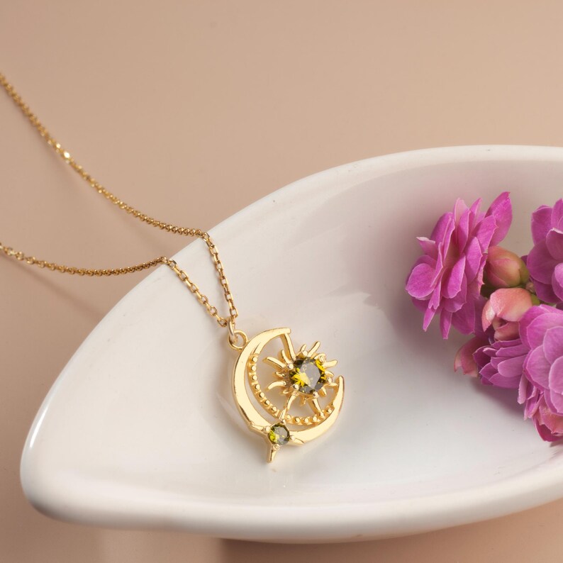 Pu&ograve; includere: Una collana dorata con un ciondolo a forma di luna e sole con una gemma verde. La collana &egrave; esposta su un piatto bianco, con fiori rosa sullo sfondo. La catena &egrave; delicata e il ciondolo presenta dettagli intricati.