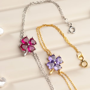 Puede incluir: Dos collares con colgantes de trébol. Uno es plateado con un trébol de piedras rosas, el otro dorado con un trébol de piedras moradas. Ambos collares tienen cadenas finas. Los colgantes tienen forma de corazón.