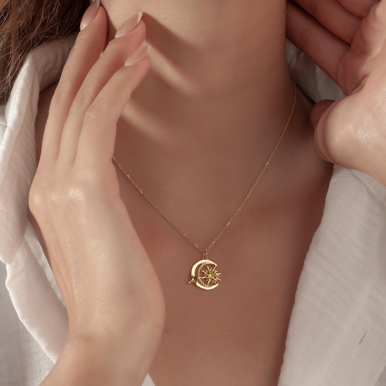 Pu&ograve; includere: Una collana dorata con un ciondolo a forma di luna crescente e un motivo a stella. Il ciondolo ha una piccola gemma verde al centro. La collana &egrave; indossata su una camicia bianca.
