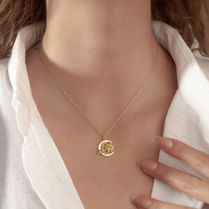 Pu&ograve; includere: Una collana dorata con un ciondolo a forma di luna crescente e sole. Il ciondolo presenta una piccola stella e un motivo a raggiera con una pietra gialla centrale. La collana &egrave; indossata su una camicia bianca.