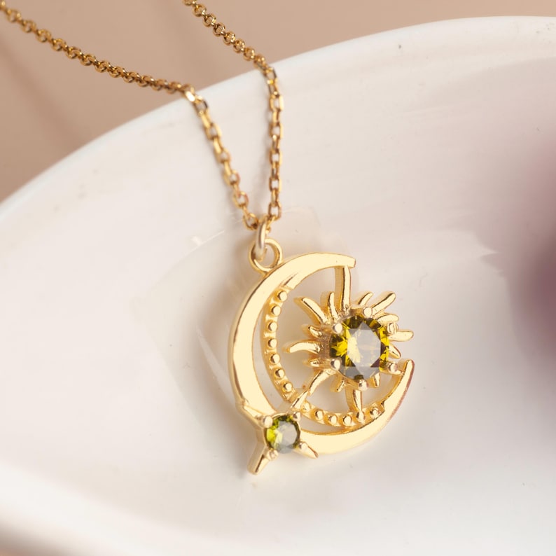 Pu&ograve; includere: Collana dorata con pendente a forma di luna e sole. Il pendente presenta una gemma verde centrale e un piccolo accento di gemma verde. La catena &egrave; delicata e la collana &egrave; esposta su una superficie bianca.