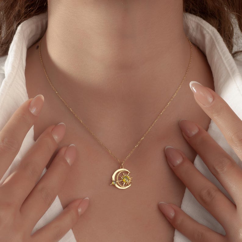 Pu&ograve; includere: Collana dorata con un ciondolo a forma di luna crescente e sole con una piccola gemma verde. La delicata catena &egrave; visibile su una camicia bianca. Il ciondolo ha una stella e una piccola gemma verde.