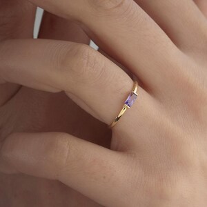Könnte beinhalten: Ein goldener Ring mit einem rechteckigen, lila Edelstein. Der Ring ist dünn und zart, wobei der Edelstein horizontal angeordnet ist. Ein schlichtes, elegantes Schmuckstück.