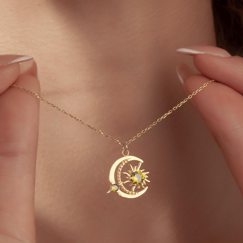 Pu&ograve; includere: Una collana dorata con un ciondolo a forma di luna crescente e sole. Il ciondolo ha pietre preziose gialle ed &egrave; sospeso a una catena delicata. La collana &egrave; indossata.