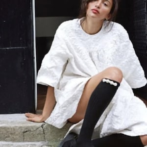 May include: A woman wearing a white, textured dress with a black knee-high sock featuring a pearl embellishment. She is wearing black leather clogs with a buckle.