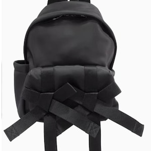 May include: Black backpack with a rounded shape, featuring a top handle and adjustable straps. The front of the backpack has decorative black straps arranged in a bow-like design. A small side pocket is visible on the left side.