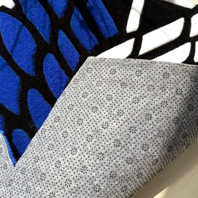 May include: A rug with a blue, black, and white geometric pattern. The rug's underside is a gray, non-slip material. The design features blue shapes outlined in black, with white shapes filling the remaining spaces. The rug is likely for home decor.