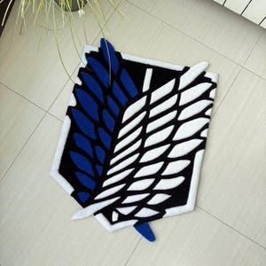 May include: A black, white, and blue rug featuring a stylized emblem design. The rug has a white border and a geometric pattern with blue and white leaf-like shapes. The rug is placed on a light-colored tiled floor.
