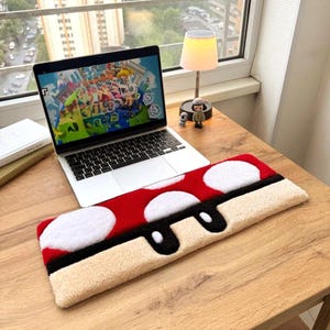 Pode incluir: Um tapete vermelho e branco em forma de cogumelo com uma borda preta está colocado em uma mesa de madeira. Um laptop exibindo um videogame está aberto ao lado do tapete. Uma pequena lâmpada e estatuetas também estão na mesa.