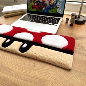 Könnte beinhalten: Eine rot-weißes Schreibtischunterlage in Pilzform mit schwarzem Rand und beigefarbener Basis. Die Matte liegt auf einem Holztisch neben einem Laptop, auf dem ein Videospiel zu sehen ist. Bücher und Figuren befinden sich ebenfalls auf dem Schreibtisch.