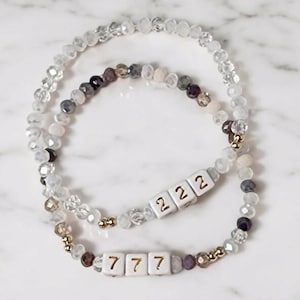 May include: Two beaded bracelets with white beads and gold accents. One bracelet has white beads with gold numbers 222 and the other has white beads with gold numbers 777.