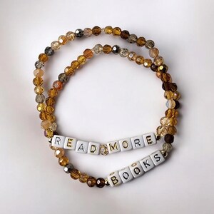 May include: Two beaded bracelets with the words "READ MORE BOOKS" spelled out in white letter beads. The bracelets are made of amber, brown, and gold-colored beads, and are displayed on a white background.