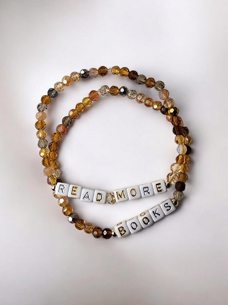 May include: Two beaded bracelets with the words "READ MORE BOOKS" spelled out in white letter beads. The bracelets are made of amber, brown, and gold-colored beads, and are displayed on a white background.