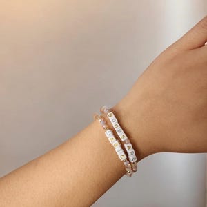 May include: Two beaded bracelets with the words "BOOKS ARE MAGIC" spelled out in white letter beads. The bracelets are on a wrist, and the beads are interspersed with small, iridescent beads.