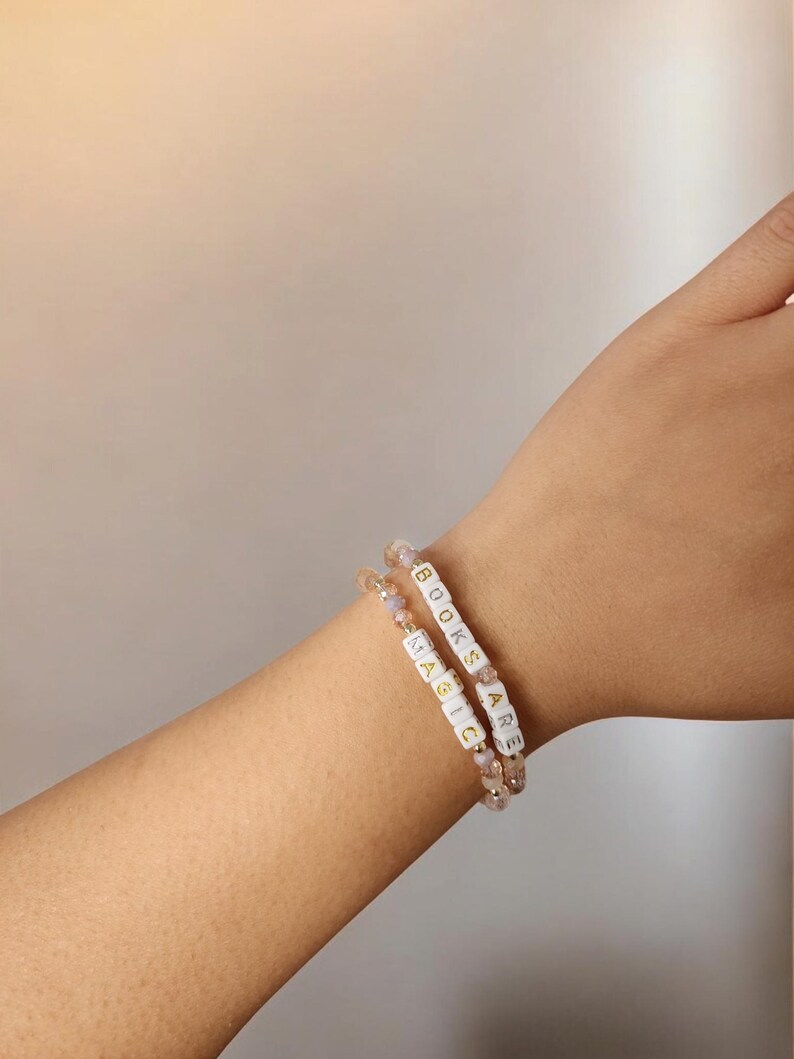 May include: Two beaded bracelets with the words "BOOKS ARE MAGIC" spelled out in white letter beads. The bracelets are on a wrist, and the beads are interspersed with small, iridescent beads.