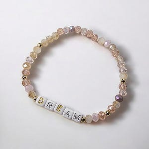 May include: A beaded bracelet with a gradient of light pink, peach, and gold beads. The bracelet has a charm with the word "DREAM" spelled out in gold letters.