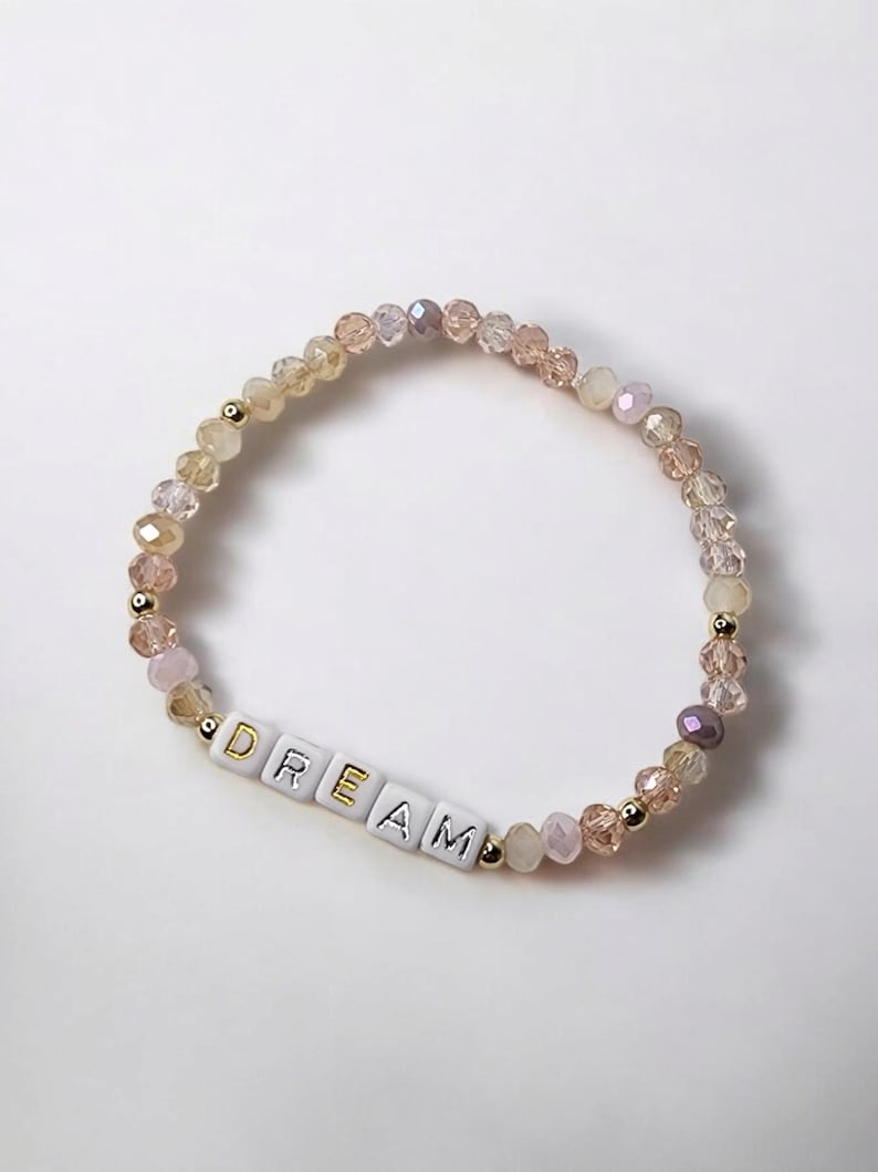 May include: A beaded bracelet with a gradient of light pink, peach, and gold beads. The bracelet has a charm with the word "DREAM" spelled out in gold letters.