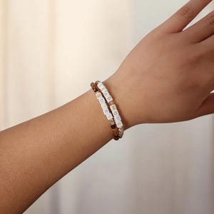 May include: Two beaded bracelets on a wrist. One bracelet has brown and amber beads, the other has white letter beads spelling "READ MORE BOOKS". The bracelets are worn together, showcasing a literary theme.