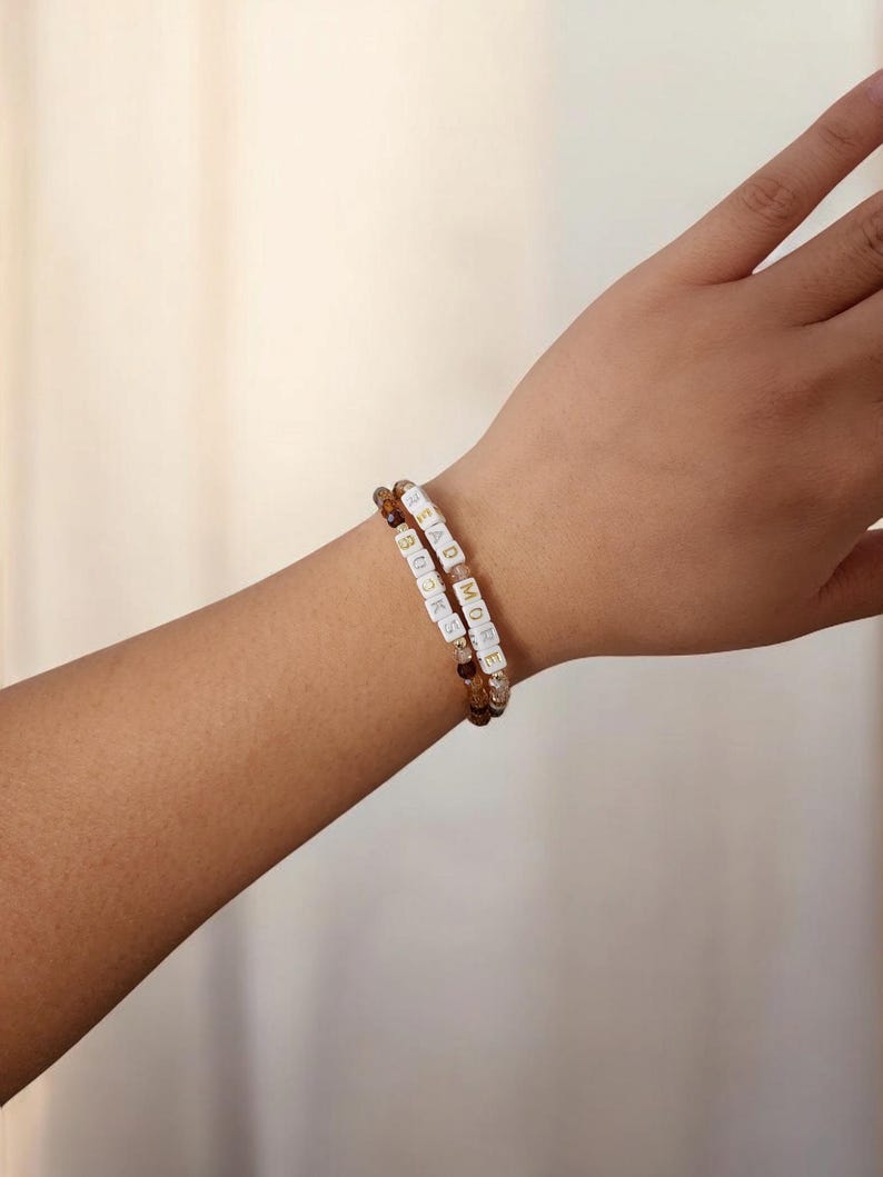 May include: Two beaded bracelets on a wrist. One bracelet has brown and amber beads, the other has white letter beads spelling "READ MORE BOOKS". The bracelets are worn together, showcasing a literary theme.