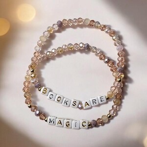 May include: Two beaded bracelets with the phrases "BOOKS ARE" and "MAGIC" spelled out in white letter beads. The bracelets are made of clear and gold-colored beads.