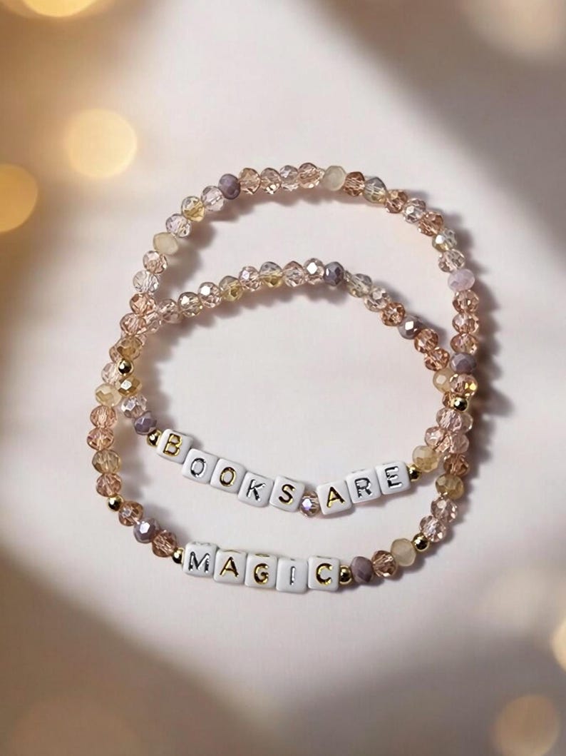 May include: Two beaded bracelets with the phrases "BOOKS ARE" and "MAGIC" spelled out in white letter beads. The bracelets are made of clear and gold-colored beads.