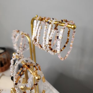 May include: A gold metal stand with multiple beaded bracelets. The bracelets are made of various colors, including white, pink, brown, and clear. Each bracelet has small white beads with gold lettering spelling out words.