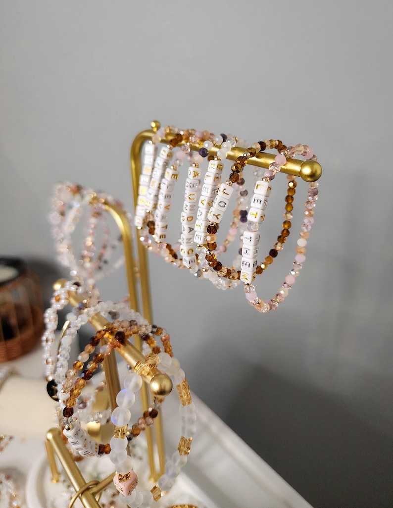 May include: A gold metal stand with multiple beaded bracelets. The bracelets are made of various colors, including white, pink, brown, and clear. Each bracelet has small white beads with gold lettering spelling out words.
