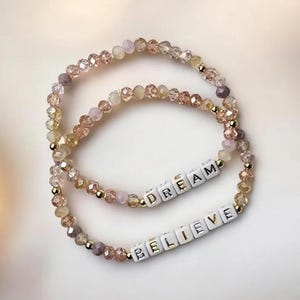 Op de afbeelding: Twee kralenarmbanden met de woorden "DREAM" en "BELIEVE" gespeld met witte letterkralen. De kralen zijn een mix van lichtroze, beige en goud.