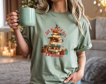 May include: Sage green t-shirt with a Christmas tree design made of books, fairy lights, and gifts. The text "All Booked for Christmas" is in red script. The shirt is a casual, festive apparel item.