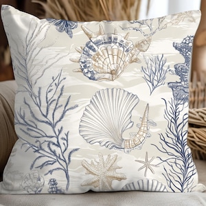 May include: Square decorative pillow with a coastal theme. The pillow features a cream-colored background with illustrations of seashells, starfish, and coral in shades of blue, beige, and white. The pillow is designed for home decor.