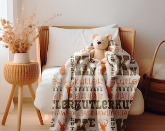 May include: A cream-colored baby blanket with a repeating pattern of deer and names in brown and orange. A teddy bear sits on the bed, partially covered by the blanket. A woven side table with a vase of dried flowers and a wicker basket are also visible.