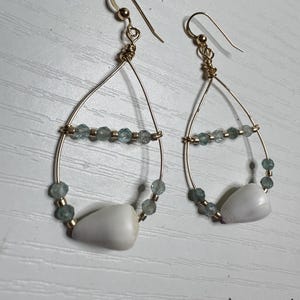 May include: A pair of gold-toned wire earrings with a teardrop design. Each earring features a white shell, blue-green beads, and a gold-colored hook. The earrings are displayed on a white surface.