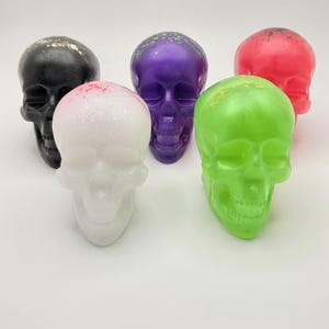 Puede incluir: Cinco figuritas de calavera de colores, cada una con un diseño diferente de pintura y salpicaduras. Las calaveras están hechas de un material liso y brillante y están dispuestas en fila sobre una superficie blanca.