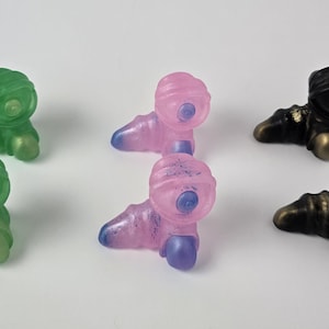 May include: Three small, hand-painted figurines in green, pink and black. Each figurine has a rounded head with a single eye and a body with three legs.