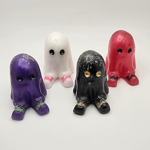 Puede incluir: Cuatro pequeñas figuritas de fantasmas, cada una de un color diferente: morado, blanco, negro y rojo. Cada fantasma tiene ojos negros y está sentado con las piernas cruzadas.