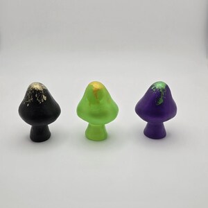 May include: Three mushroom-shaped ornaments. One is black with gold detailing, the second is bright green with yellow accents, and the third is purple with green detailing. Each has a rounded cap and a stem.