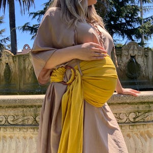 May include: A pregnant person wearing a long, light brown dress with a yellow fabric sash tied around their waist. The sash is tied in a knot at the front and has a large, round, metal ring.