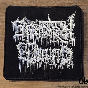 May include: A square black fabric patch featuring white embroidered text in a stylised, dripping, jagged font. The embroidery creates a dense, textured appearance against the solid black background, with clean, stitched edges forming a border.