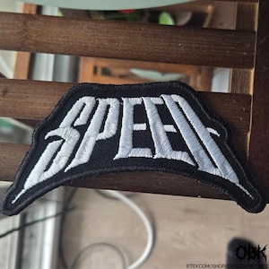 May include: A black embroidered patch featuring the word SPEED in large, white, stylised capital letters. The patch has a distinct, angular, and elongated shape with a thick black border surrounding the white stitched lettering.