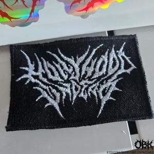 May include: Black rectangular patch with white embroidered text. The text appears to be stylized and gothic in design. The patch is likely for clothing or accessories. A holographic sticker is visible in the background.
