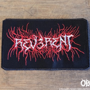 Puede incluir: Un parche rectangular bordado en negro con el texto blanco "REV3RENT" centrado en el medio. Las letras están rodeadas por un intrincado bordado de hilo rojo que se extiende y se asemeja a estructuras orgánicas como raíces o venas.