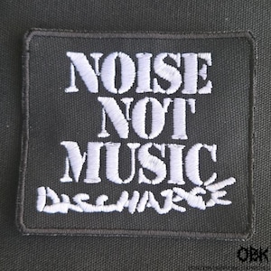 May include: Black rectangular patch with white embroidered text that reads "NOISE NOT MUSIC" above "DISCHARGE". The patch has a black border and is likely for clothing or accessories.
