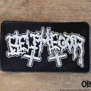May include: A rectangular black embroidered patch featuring white stylized lettering that reads Belphegor, accompanied by two white cross symbols positioned beneath the text. The patch has a black stitched border and a textured fabric base.
