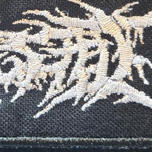 May include: A black fabric patch featuring intricate, dense white embroidery in a jagged, abstract, and organic pattern. The stitching creates a textured, raised effect against the dark background, with sharp, branching lines that resemble roots or vines.