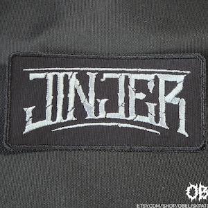Puede incluir: Un parche rectangular negro bordado que presenta la palabra JINJER cosida en una fuente gris claro estilizada y desgastada. El parche tiene un borde negro sólido y está colocado sobre un fondo de tela negra, con un pequeño logo.