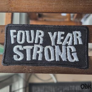 May include: A rectangular embroidered patch with a black background and white stitched lettering that reads FOUR YEAR STRONG. The patch features a thick black border and a textured fabric surface with clear, bold, capitalised text throughout.