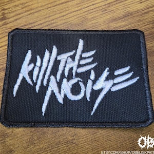 May include: Black rectangular patch with white embroidered text that reads "KILL THE NOISE". The patch has a black border and is likely an iron-on or sew-on patch. The patch is on a wooden surface.